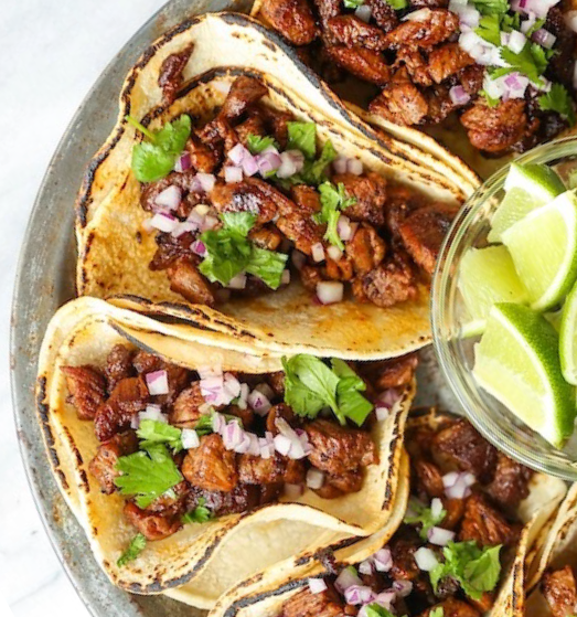 two tacos filled with a carne asada mixture next to a bowl of lemon wedges