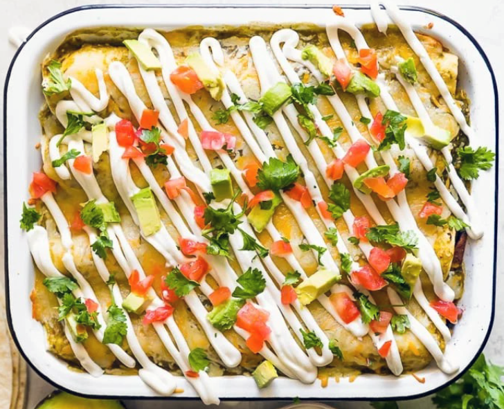 chicken, black bean and sweet potato enchiladas shown packed in a baking dish and garnished with sour cream, tomato and coriander