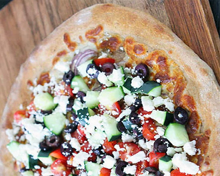 green pizza topped with olives, tomatoes, cucumber and feta cheese on a chopping board