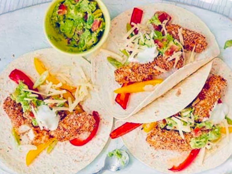 lightly-spiced breaded fish on flour tortillas with bell peppers, guacamole and sour cream next to a bowl of guacamole