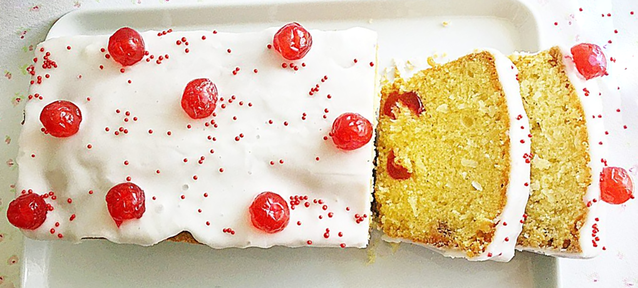 Iced glace cherry loaf cake topped with cherries served on a plate with two slices cut to reveal the centre