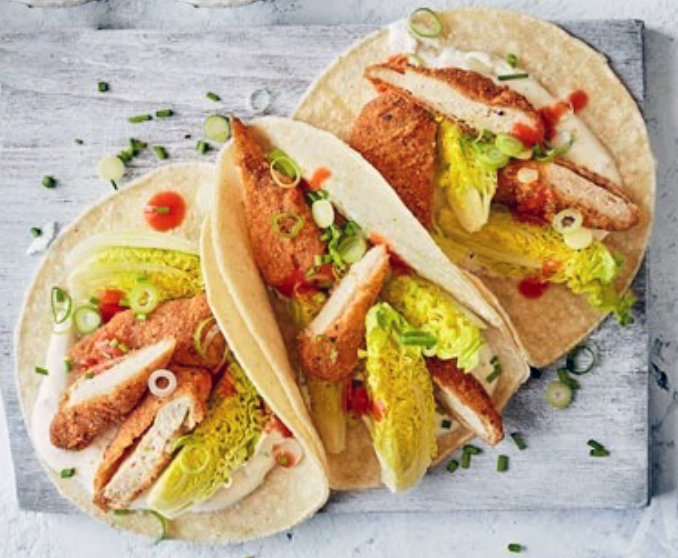 Three flour tortillas topped with sliced southern-fried chicken, lettuce and buffalo hot sauce on a white chopping board