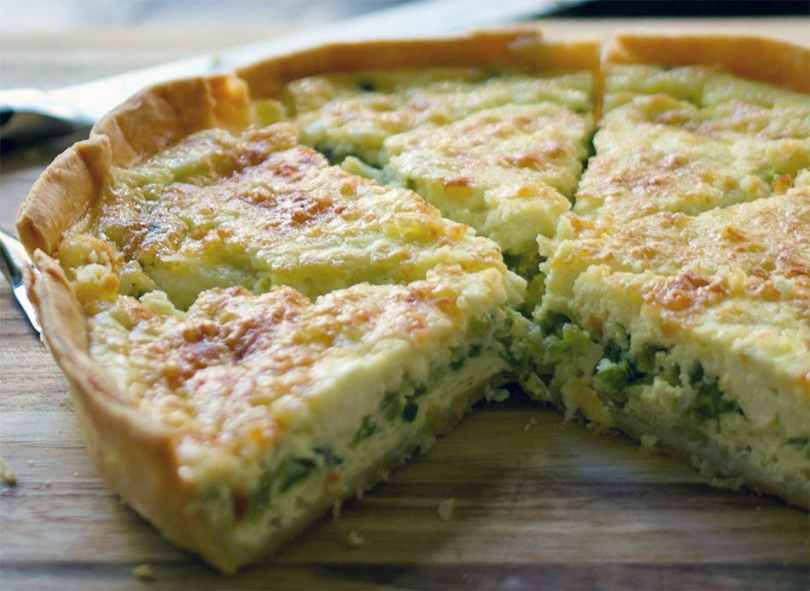 cheese and three-cornered leek quiche on a chopping board with a wedge removed to reveal the filling