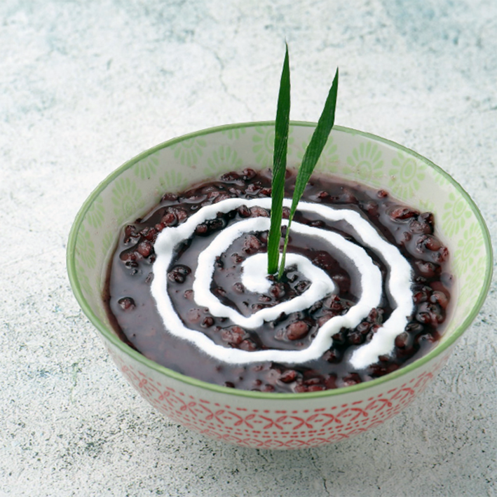 bubur pulut, Malay black rice porridge served in a bowl, garnished with a swirl of coconut milk and two slivers of pandan leaf