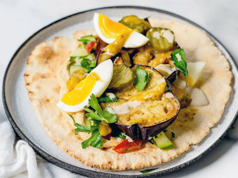 pita flatbread topped with aubergine slices, egg quarters, tahini and pickles to form a sabich