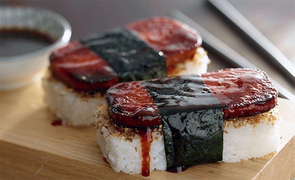 spam masubi on a chopping block with a dish of sweet ginger sauce